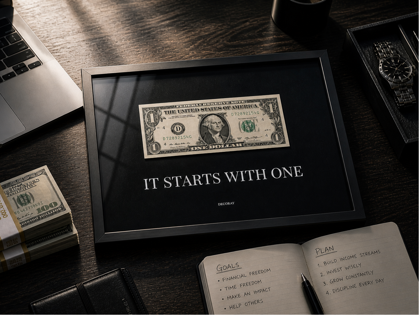 Framed one-dollar bill with 'It Starts with One' text on a desk with money and a laptop.